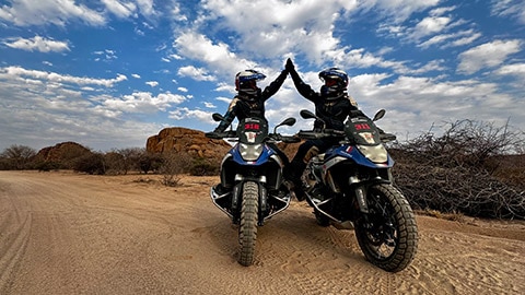 Dwie zawodniczki z francuskiej drużyny kobiet siedzą obok siebie na motocyklach R 1300 GS Competition i przybijają sobie piątkę.