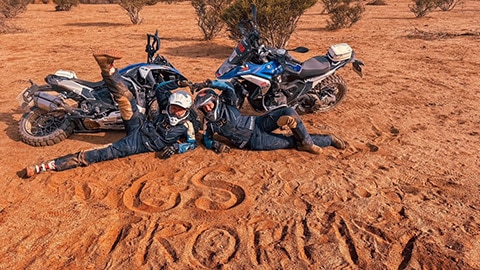 Dwie zawodniczki międzynarodowego zespołu kobiet leżą przed swoimi motocyklami na piasku, na którym utworzyły napis GS Trophy.